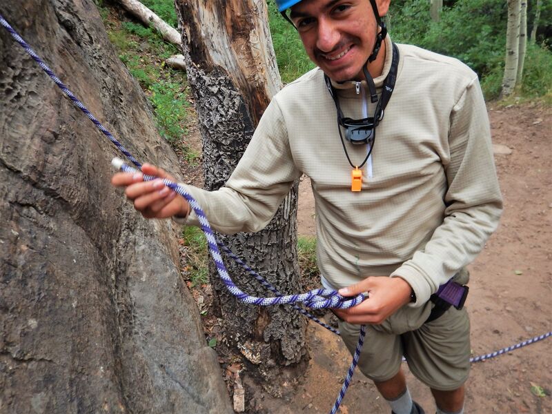 A man wearing a helmet and climbing gear is holding a rope. He is standing next to a rock face and appears to be preparing to climb. He is smiling and looking at the camera. The background shows a wooded area with a dirt path.